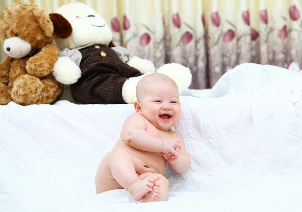 Cute smiling baby sitting on a white blanket with teddy bears in the background.